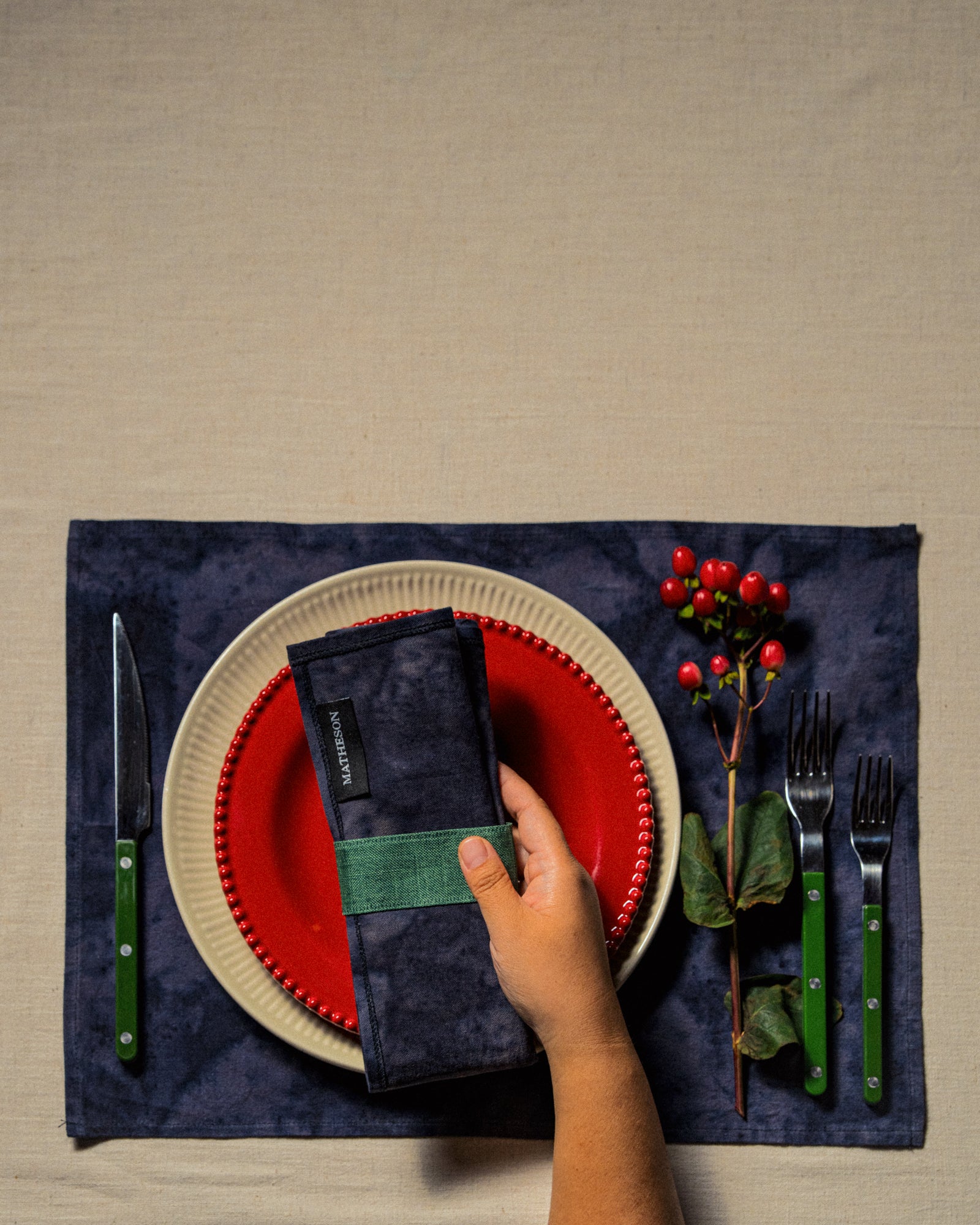 Hand setting a place at a table with a red plate, black napkin, and green cutlery on a beige surface.